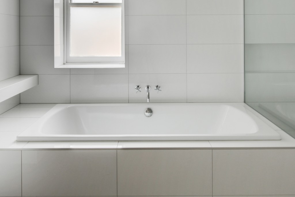 A stark, white tile bathroom with a tub and window