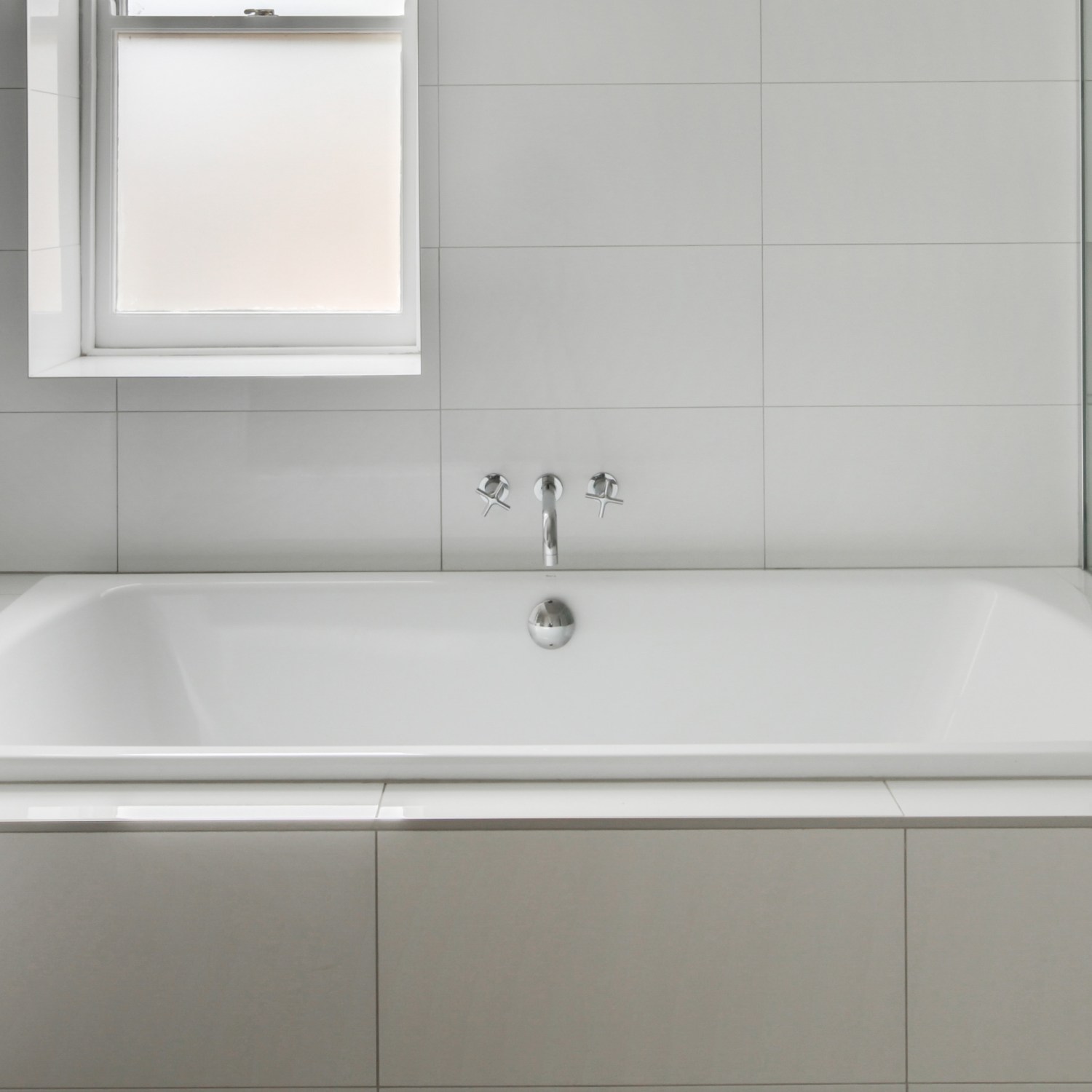 A stark, white tile bathroom with a tub and window