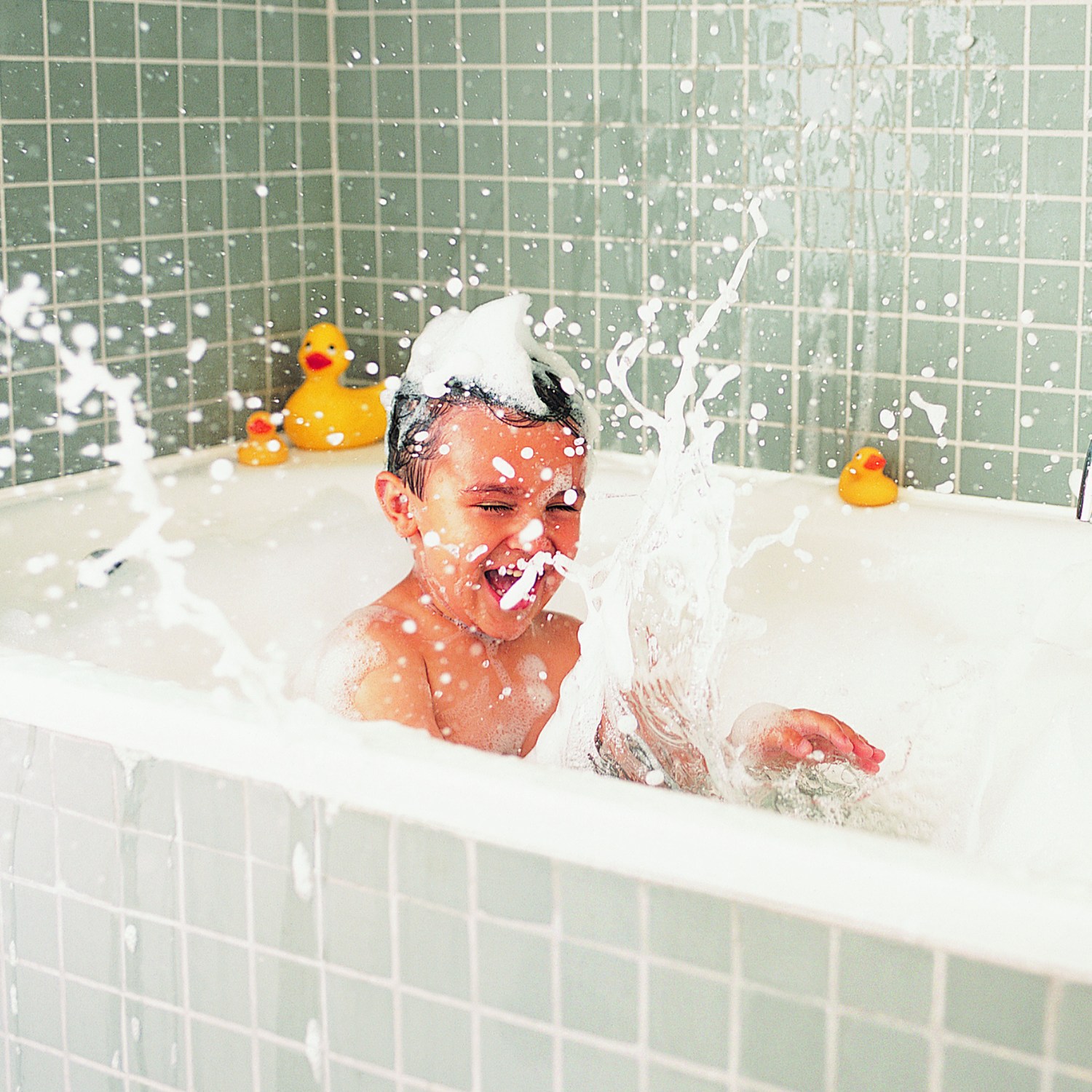 A young boy in a bath tub surrounded with bubbles & ducks