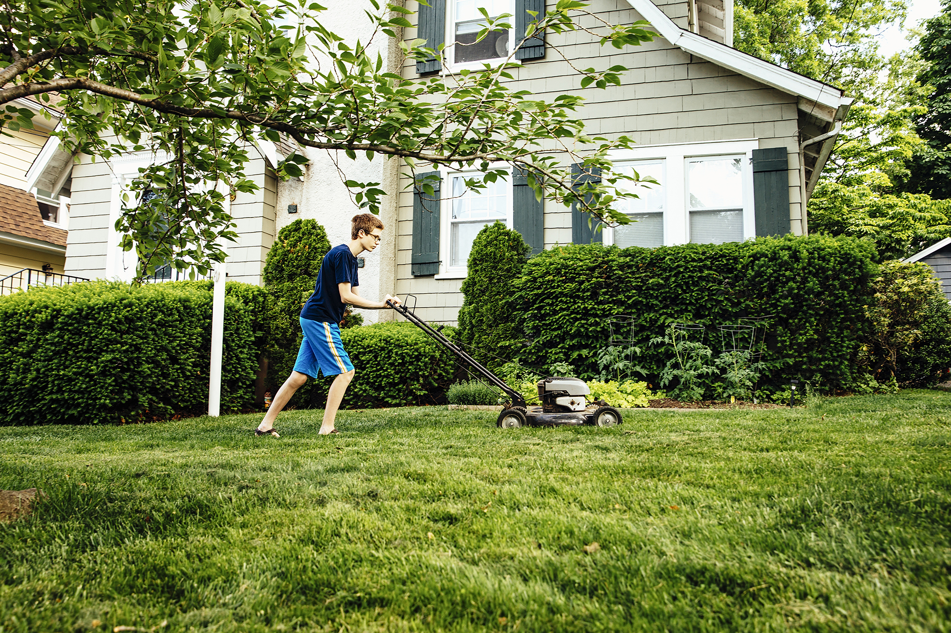 Tween boy in blue mowing lawn outside light green house