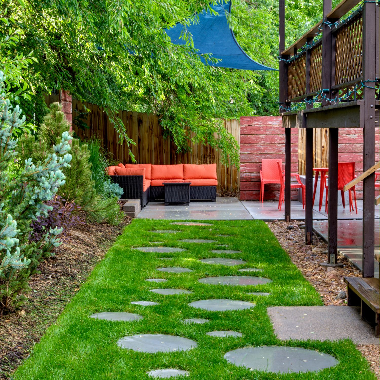 Bug free backyard patio has couch orange chairs blue shade sails a deck with paver walkway in grass