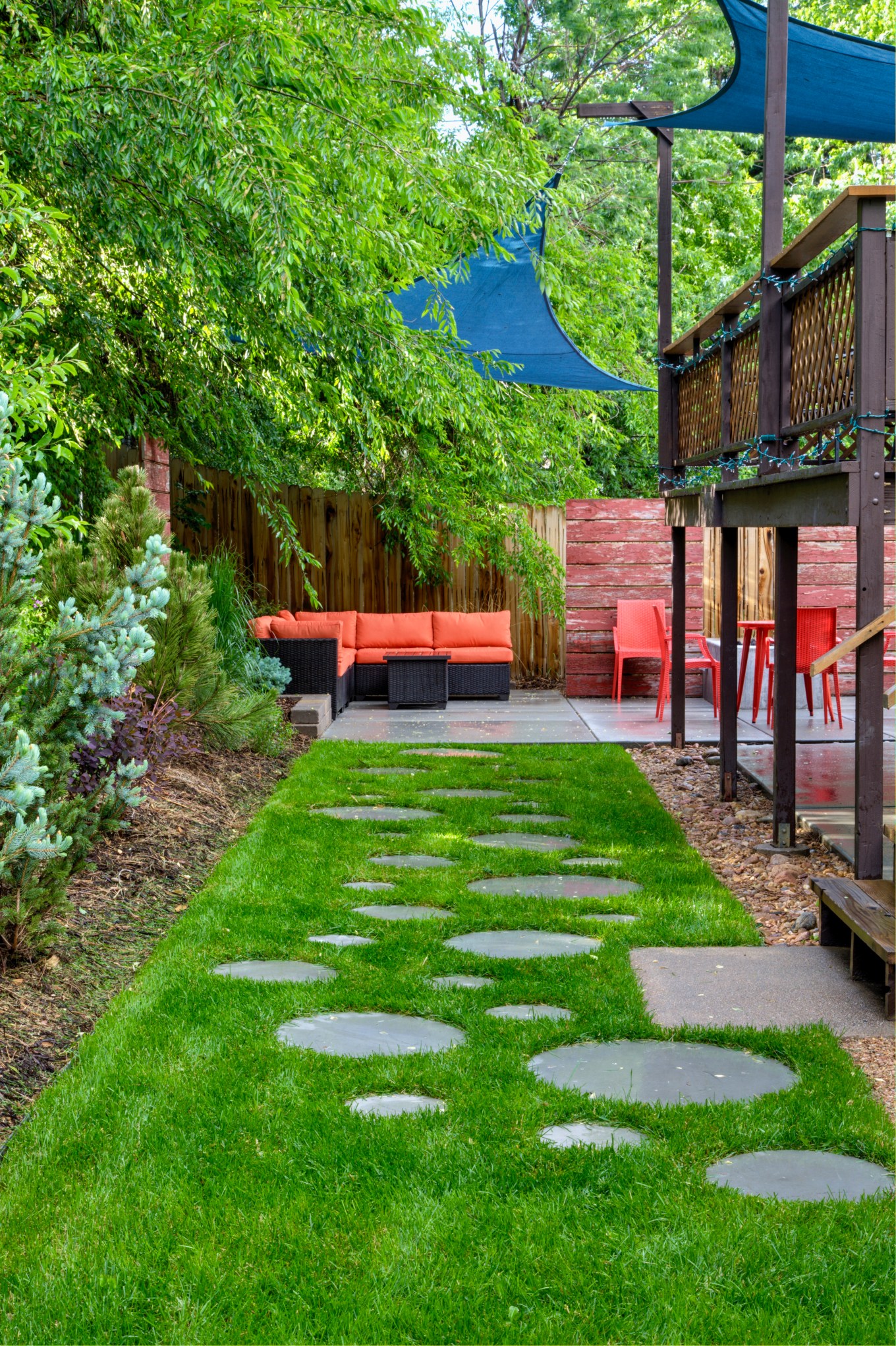 Bug free backyard patio has couch orange chairs blue shade sails a deck with paver walkway in grass