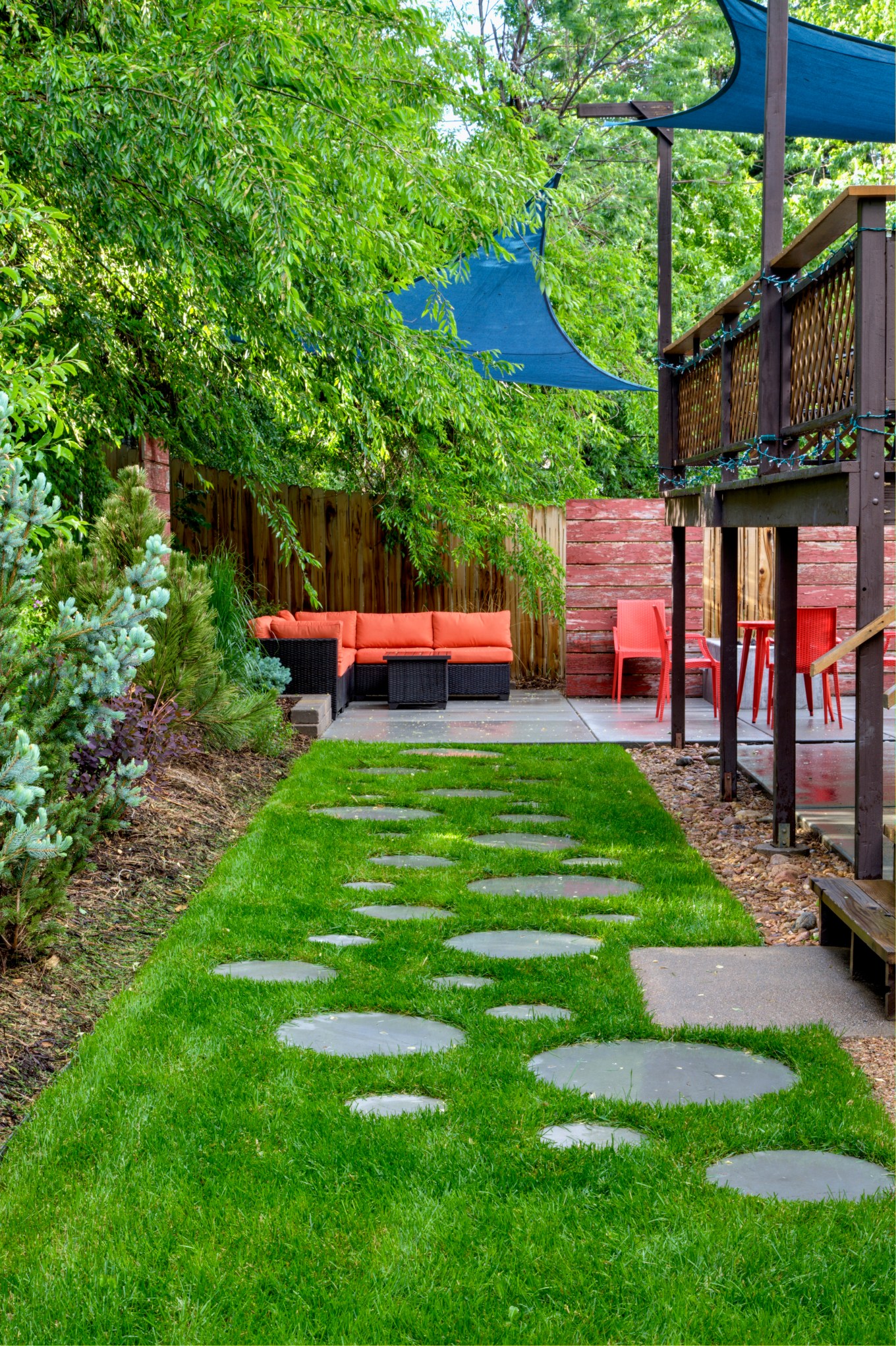 Bug free backyard patio has couch orange chairs blue shade sails a deck with paver walkway in grass