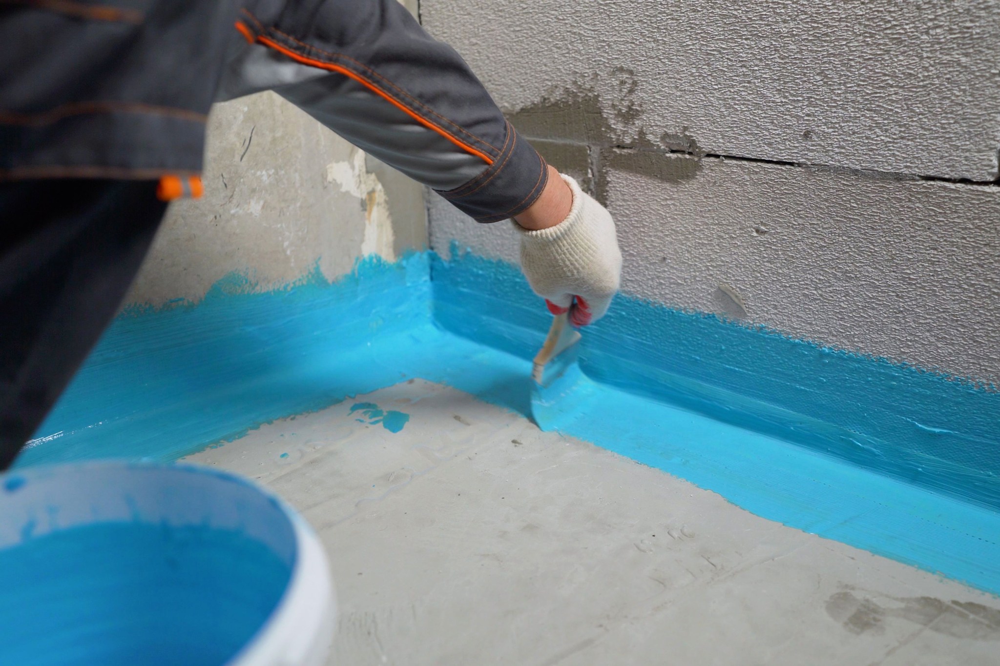 DIY waterproofing a basement with blue sealant being applied along cement floor and cinder block wall