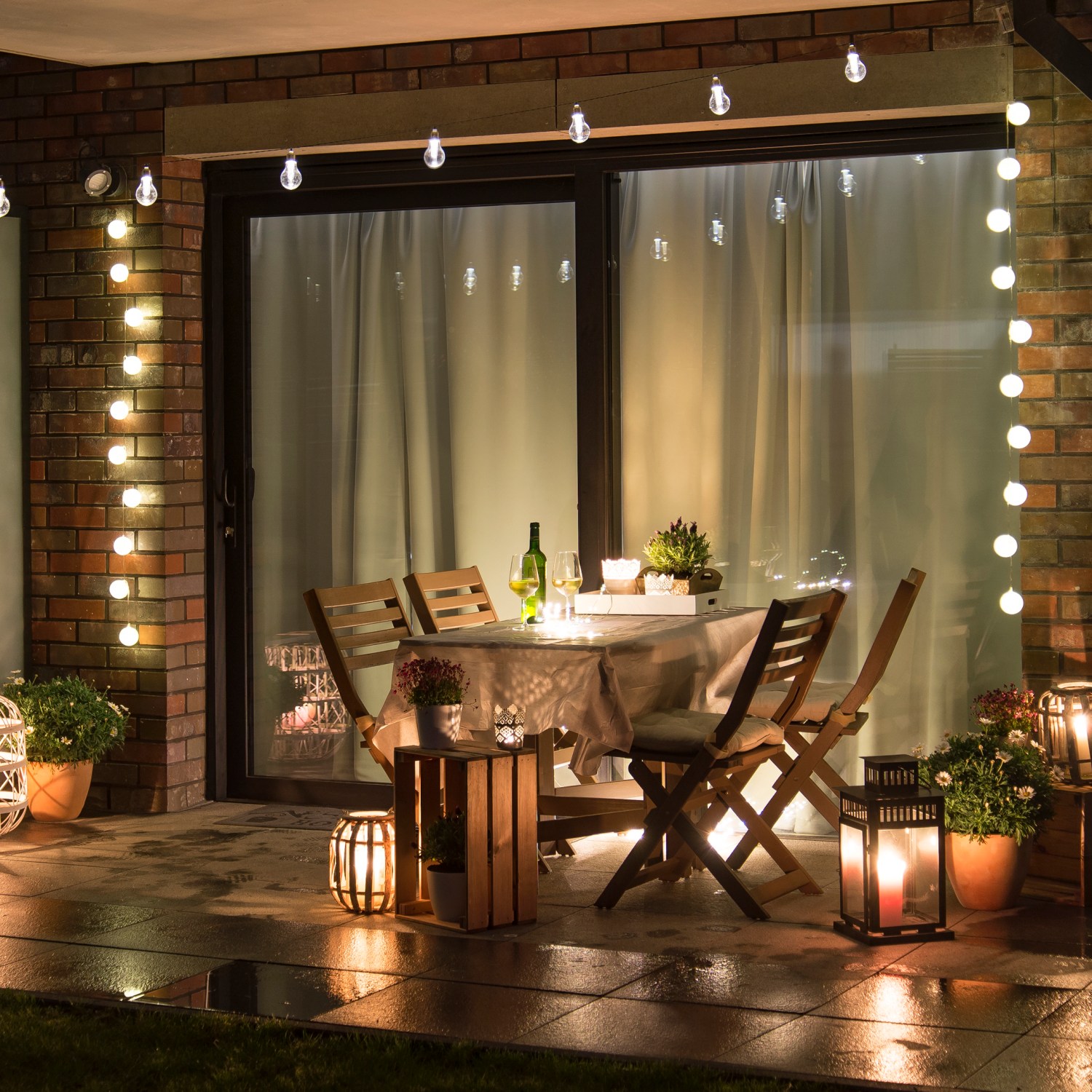 outdoor patio at night with a string lights around the sliding door and above a wooden table with candles and lanterns illuminating the terrace