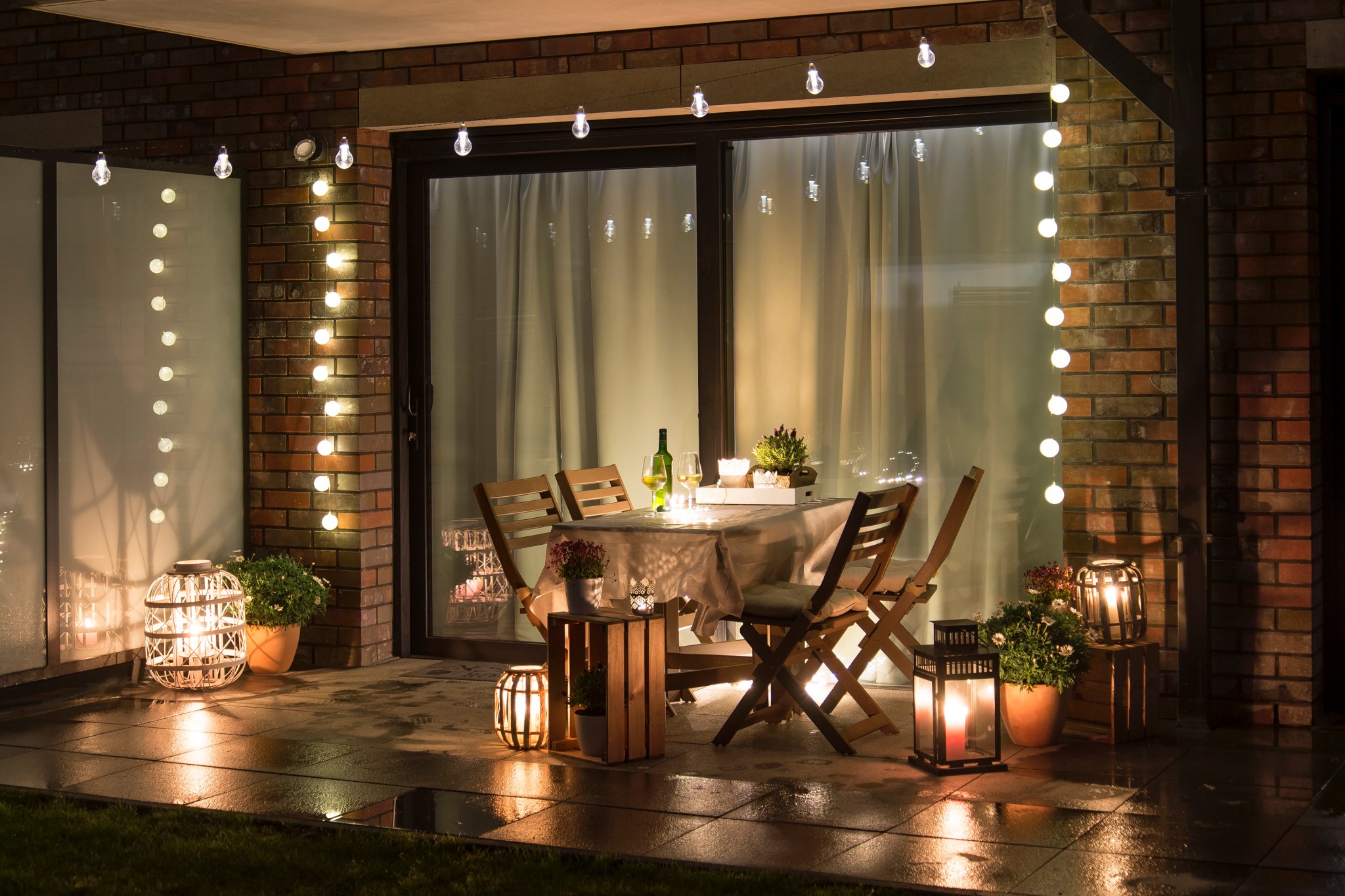 outdoor patio at night with a string lights around the sliding door and above a wooden table with candles and lanterns illuminating the terrace