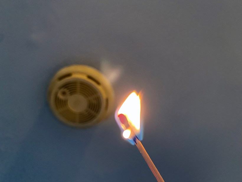 home maintenance myths that cost money a lit burning match held up to an outdated smoke detector