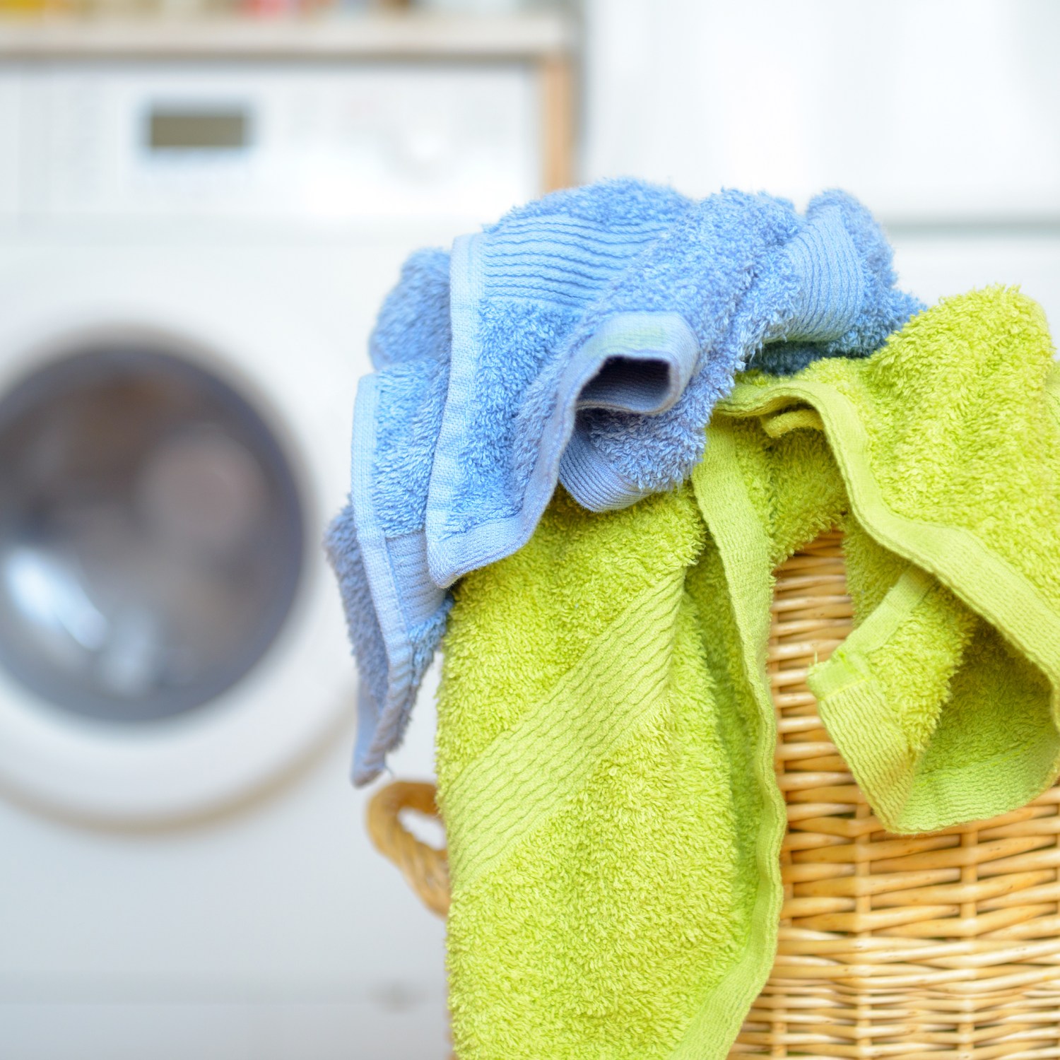 wicker laundry basket with dirty blue and green towels waiting for laundry with white washing machine in background