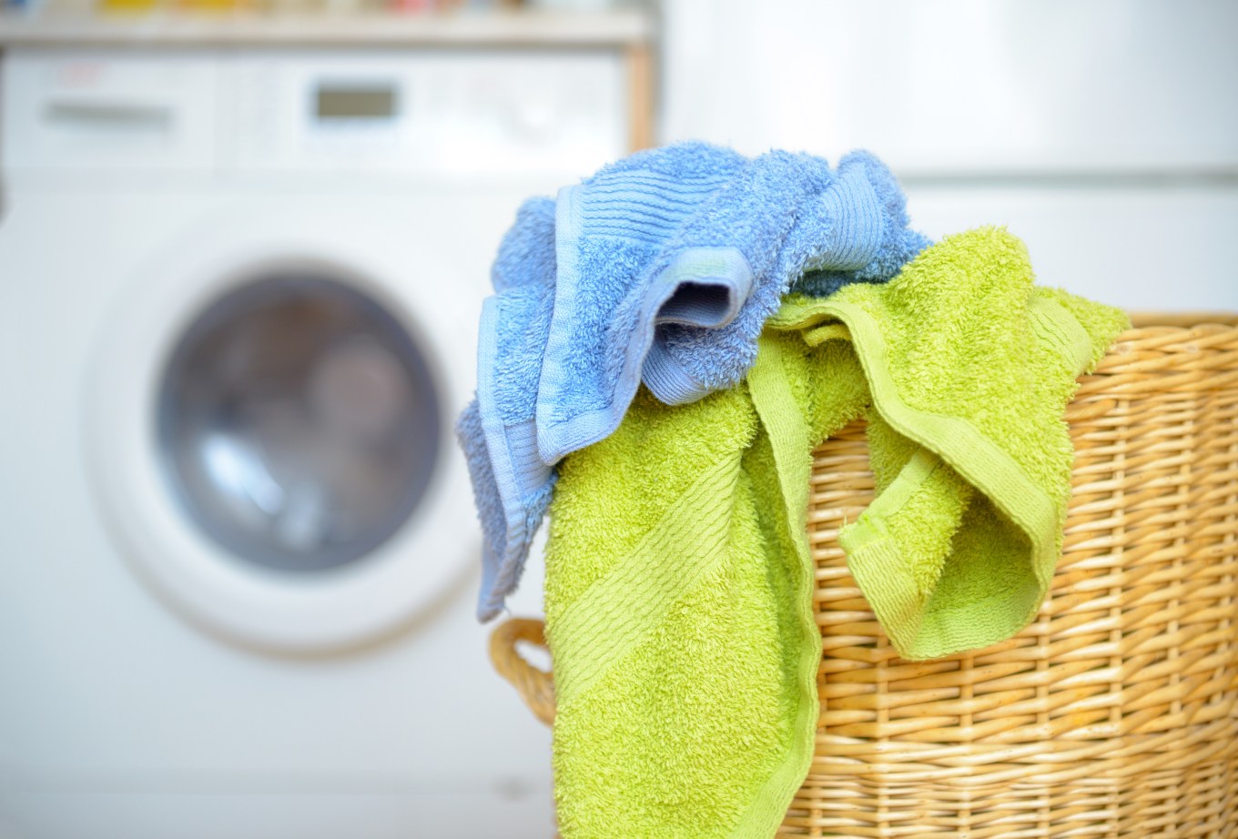 wicker laundry basket with dirty blue and green towels waiting for laundry with white washing machine in background