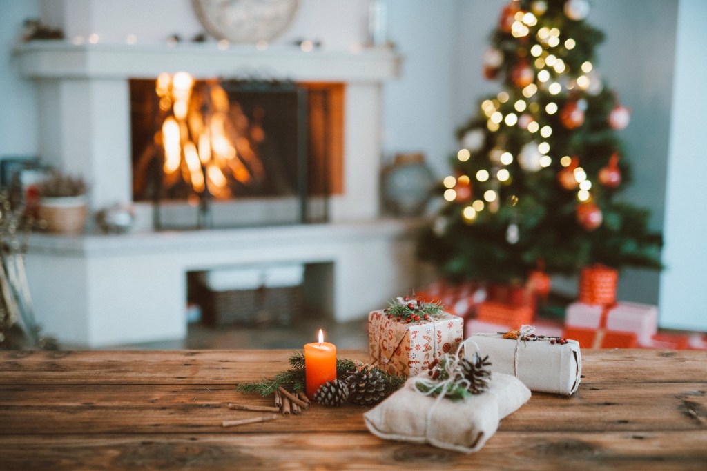 easy holiday decoration organization hacks candle and pine cones on a rustic wood table with small packages with a Christmas tree and a white fireplace with a roaring fire in the background