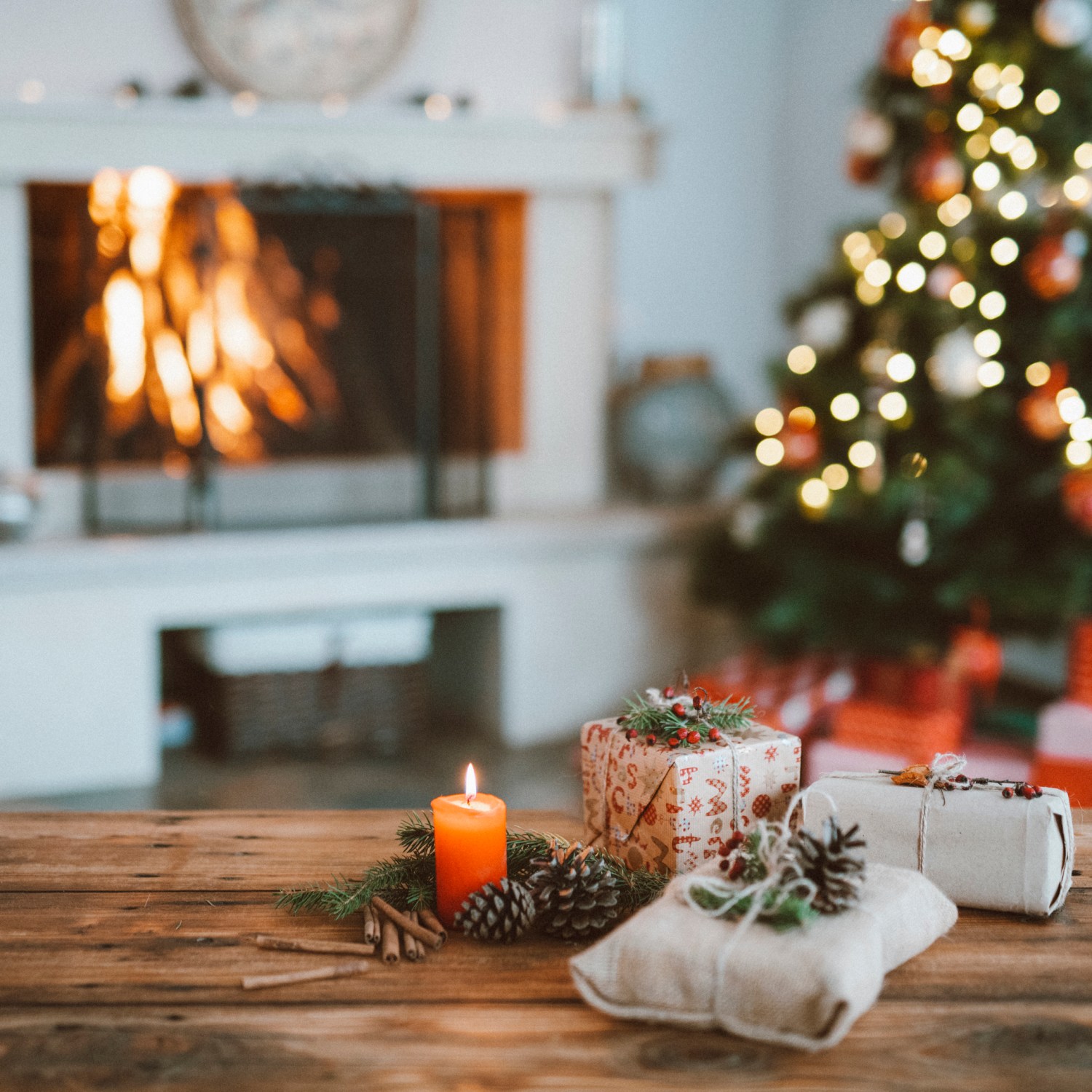 Easy holiday decoration organization hacks candle and pine cones on a rustic wood table with small packages with a Christmas tree and a white fireplace with a roaring fire in the background.