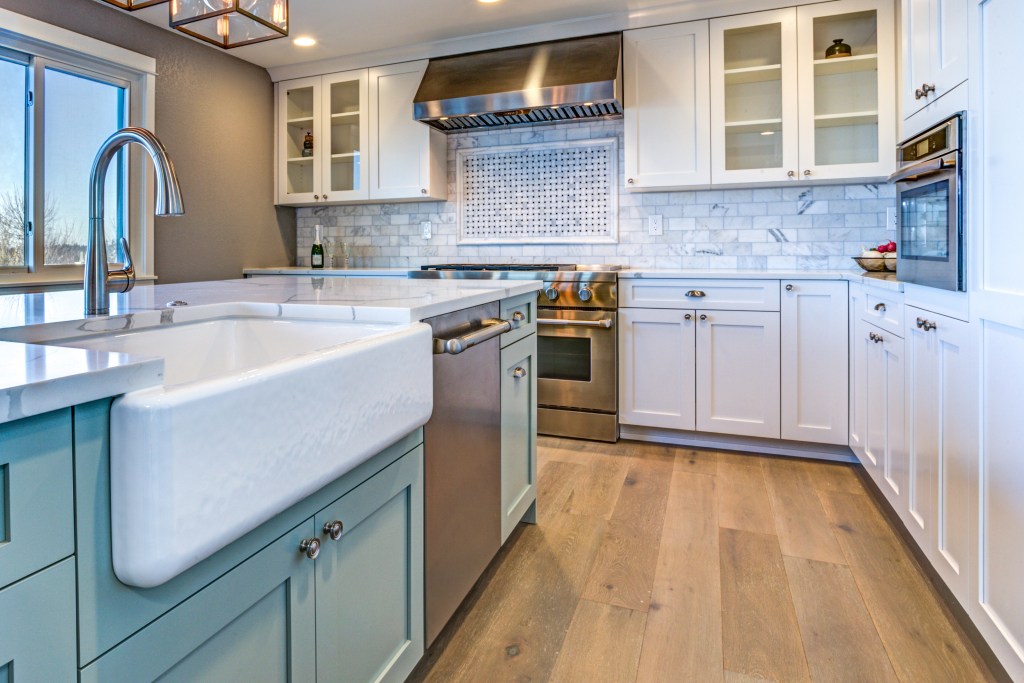 updated kitchen with light green island and white farmhouse sink with white cabinetry and white and gray pattern subway tile with a wide plank wood floor and stainless steel appliances