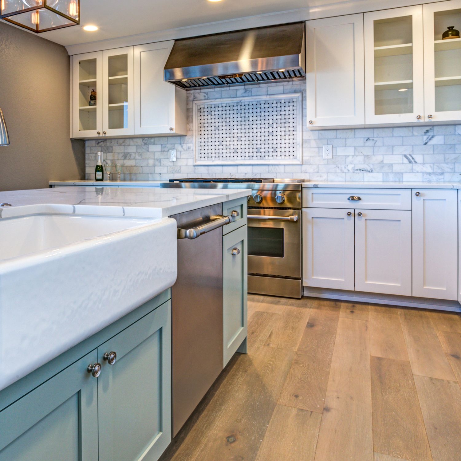 updated kitchen with light green island and white farmhouse sink with white cabinetry and white and gray pattern subway tile with a wide plank wood floor and stainless steel appliances