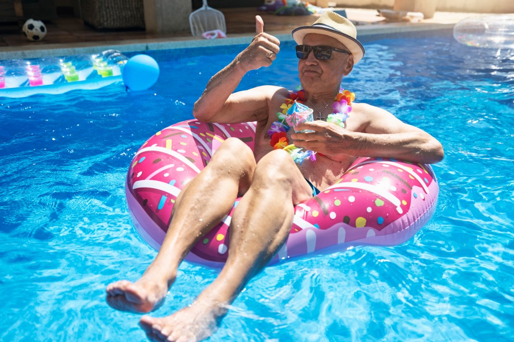 older man wearing a hat and sunglasses relaxing in pink donut floatie in a swimming pool drinking juice cocktails while giving a thumbs up