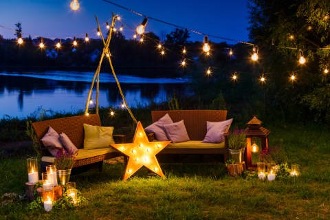 cozy outdoor space at night with two sofas with pillows and string lights and a star lamp a lantern and candles with a lake in the background
