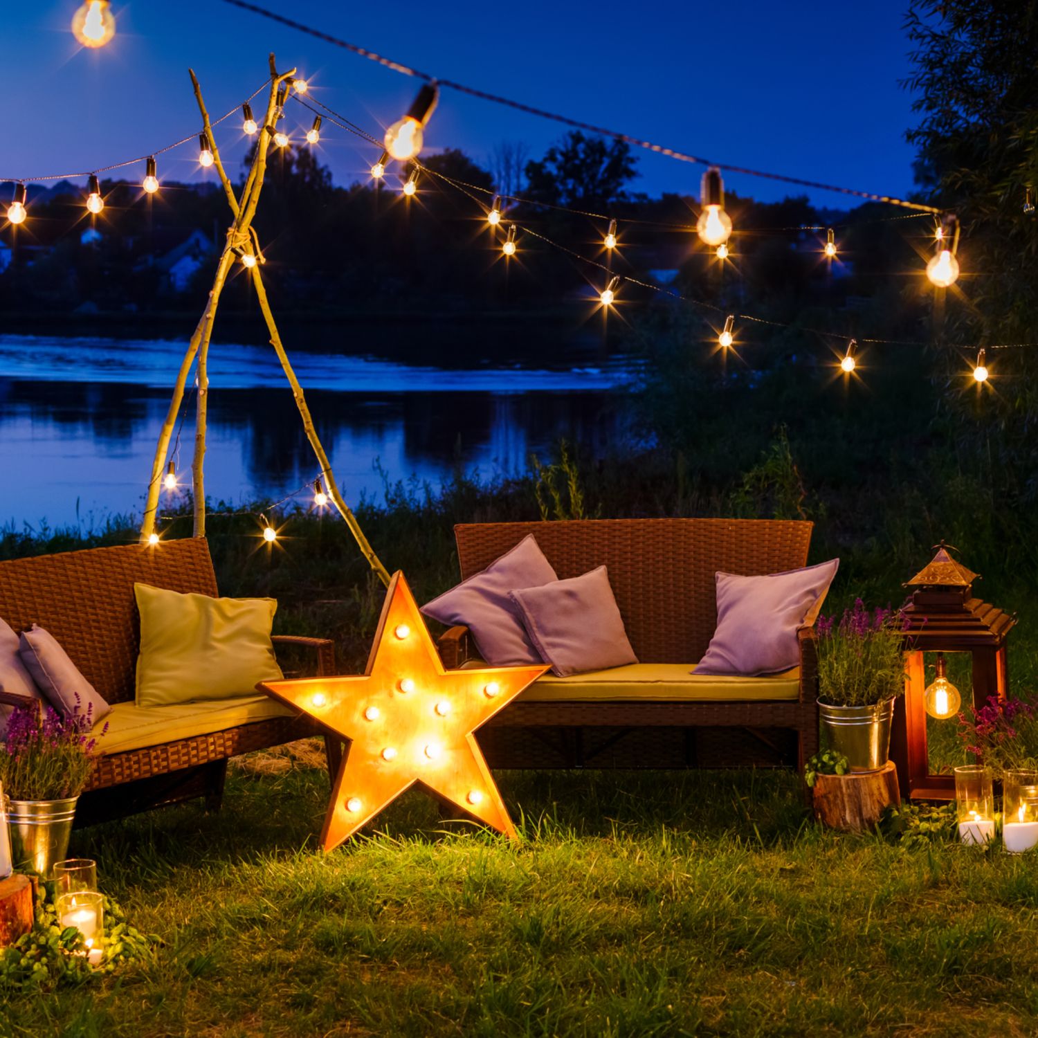 cozy outdoor space at night with two sofas with pillows and string lights and a star lamp a lantern and candles with a lake in the background