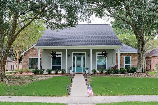 front view of renovated Acadian style home with painted and brick exterior and colorful front door with columns and tall trees in the yard