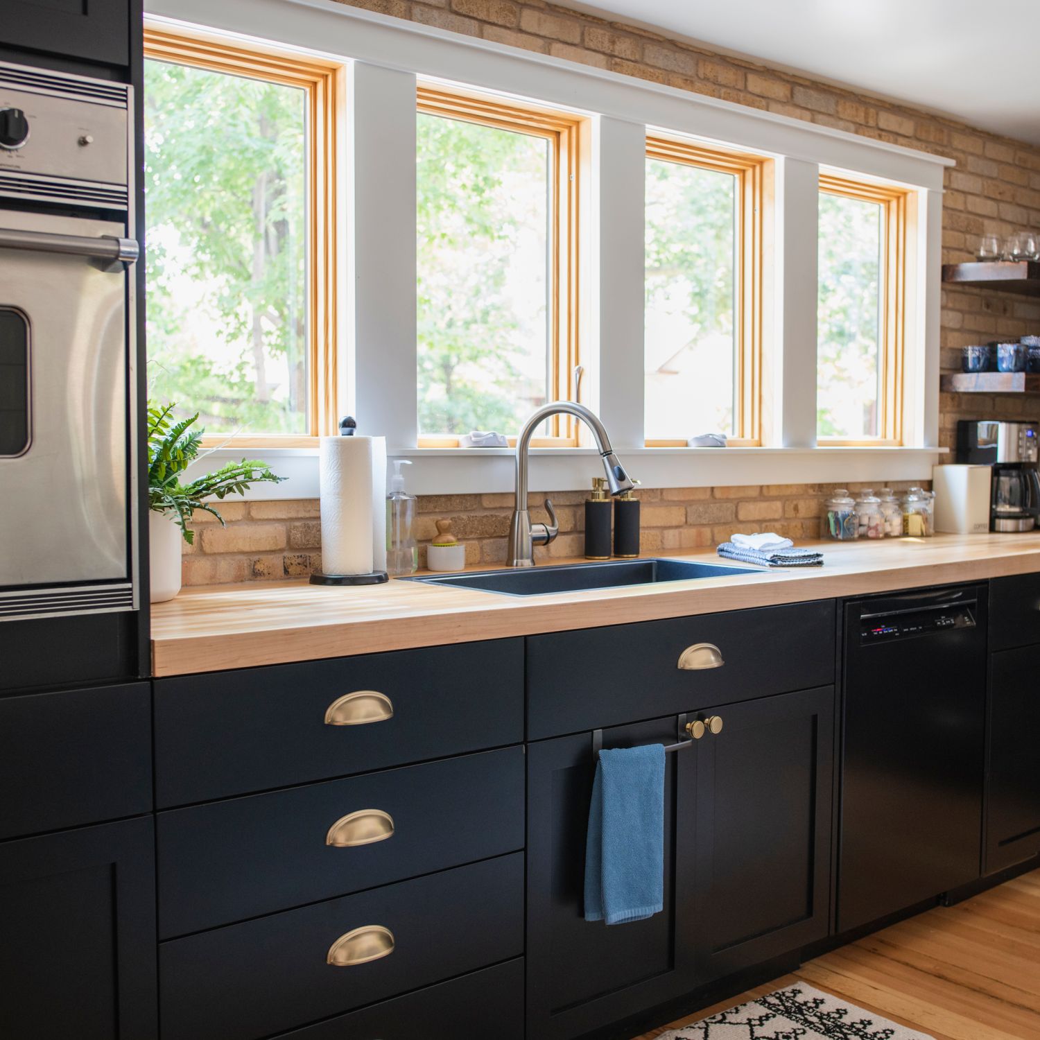 view of a kitchen with dark lower cabinets with brass hardware and brick walls with open shelving and a built in stainless steel oven with daylight shining in bright windows