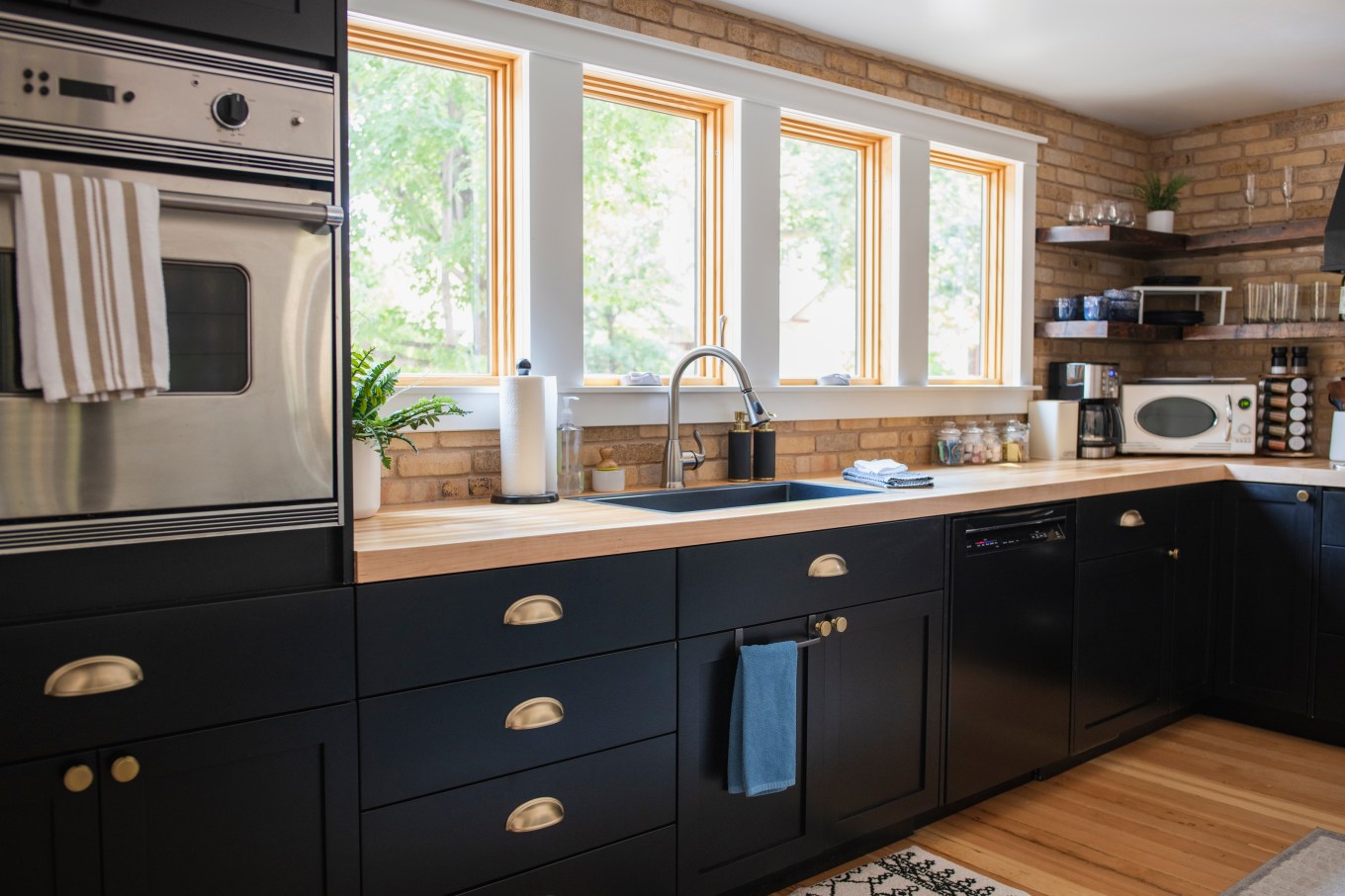 view of a kitchen with dark lower cabinets with brass hardware and brick walls with open shelving and a built in stainless steel oven with daylight shining in bright windows