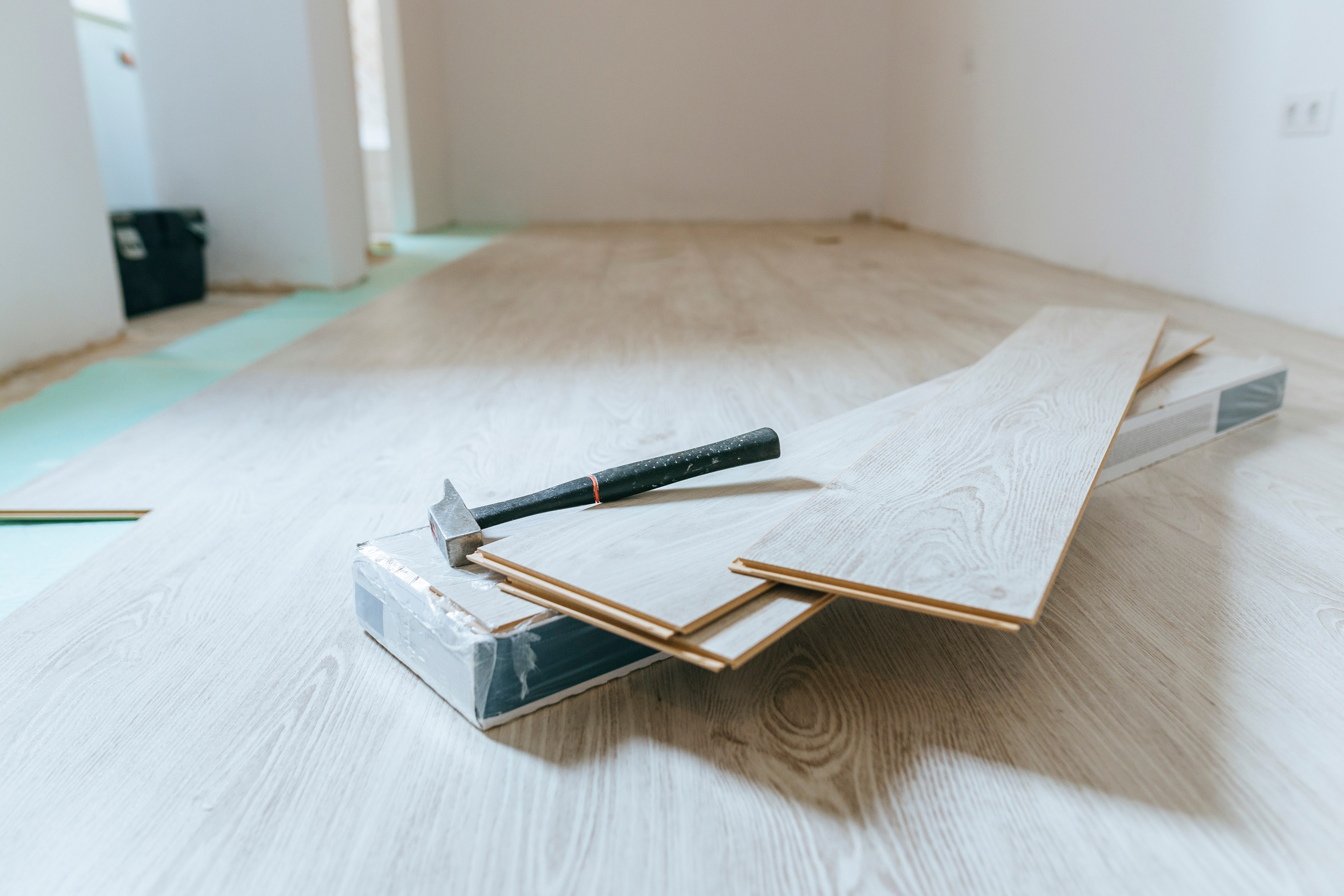 a hammer sits on sheets of wood laminate floor in an empty white room partially finished with blonde color wood grain laminate flooring