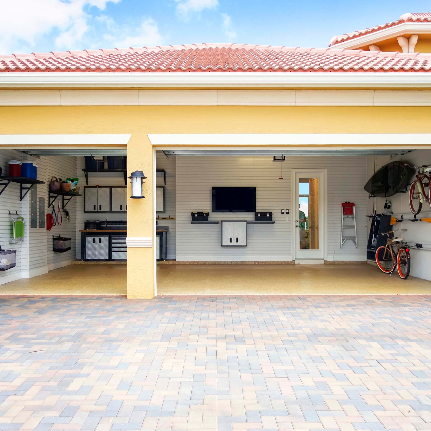 three car garage attached to a home that is well organized with shelves, bicycle racks work area and storage cabinets. There is a flat screen tv on the wall. there are no cars in the garage.