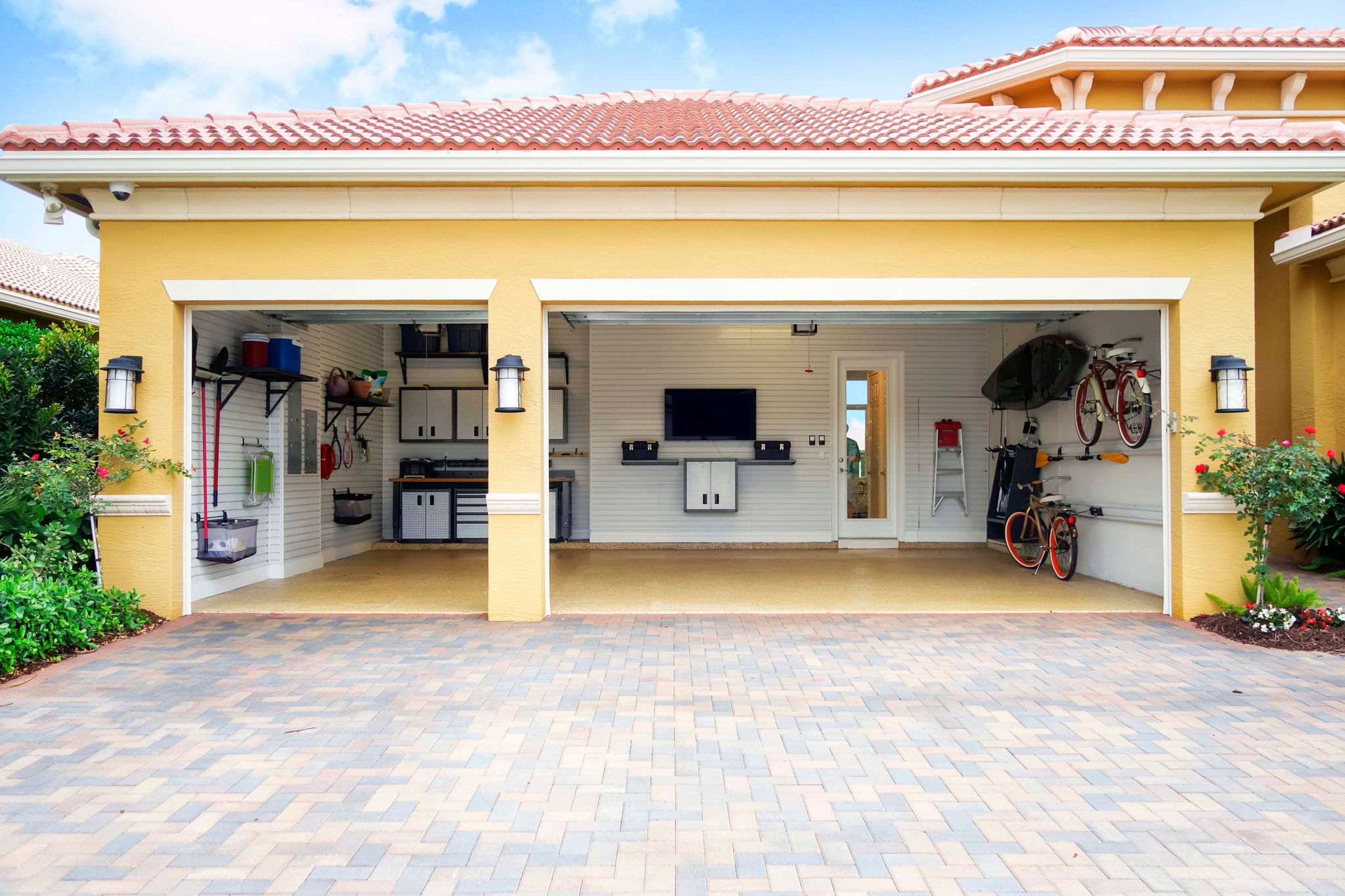 three car garage attached to a home that is well organized with shelves, bicycle racks work area and storage cabinets. There is a flat screen tv on the wall. there are no cars in the garage.