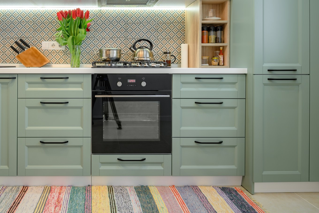 modern kitchen with light green cabinets and white countertop decorated with flowers and cooking utensils and colorful patterned backsplash with a gas cooktop and black oven