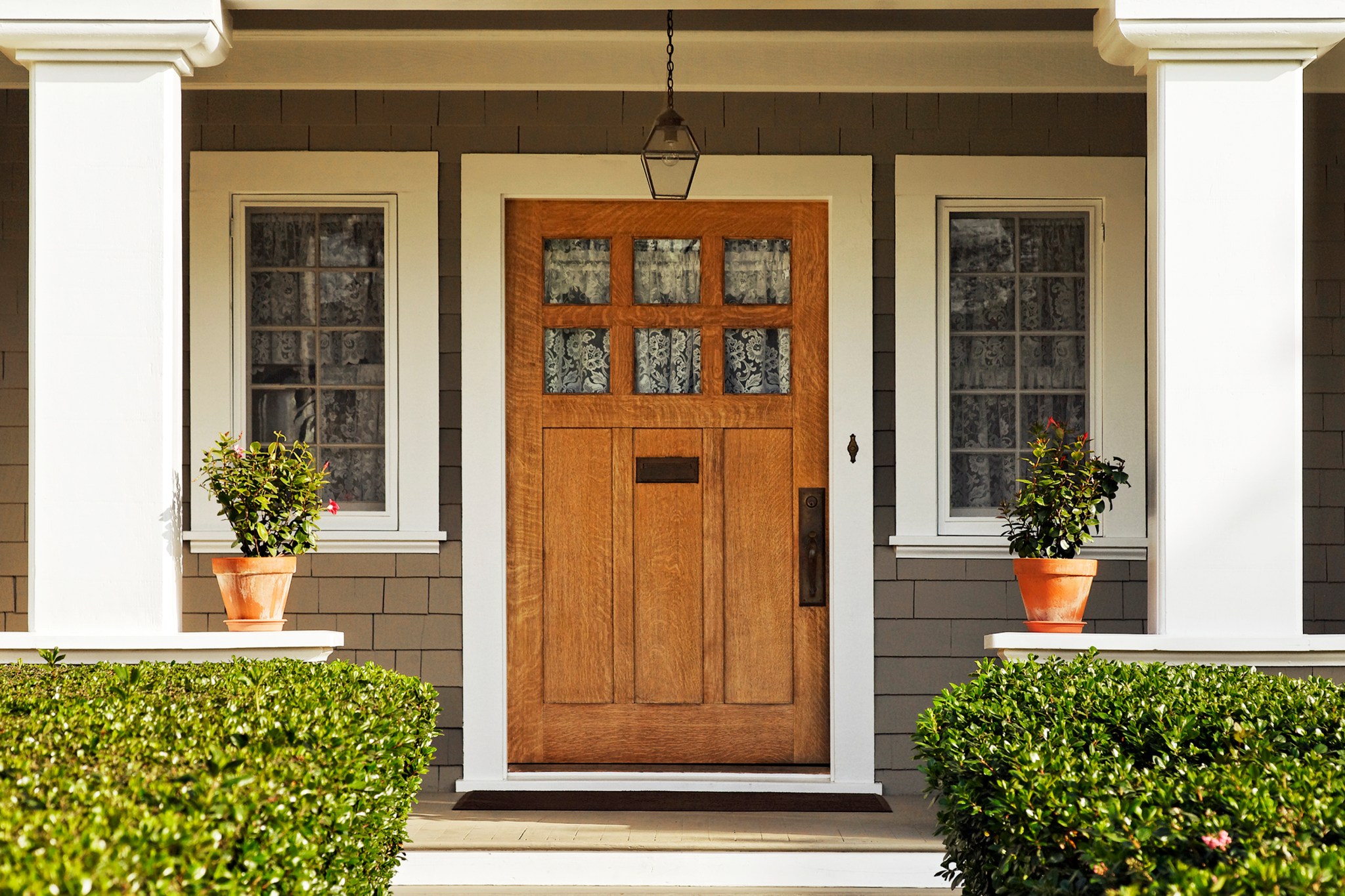 A concrete walkway bordered with hedged shrubs leads to the wooden front door of a craftsman style home with taupe shaker siding and large white columns and windows on either side of the door