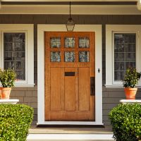 A concrete walkway bordered with hedged shrubs leads to the wooden front door of a craftsman style home with taupe shaker siding and large white columns and windows on either side of the door