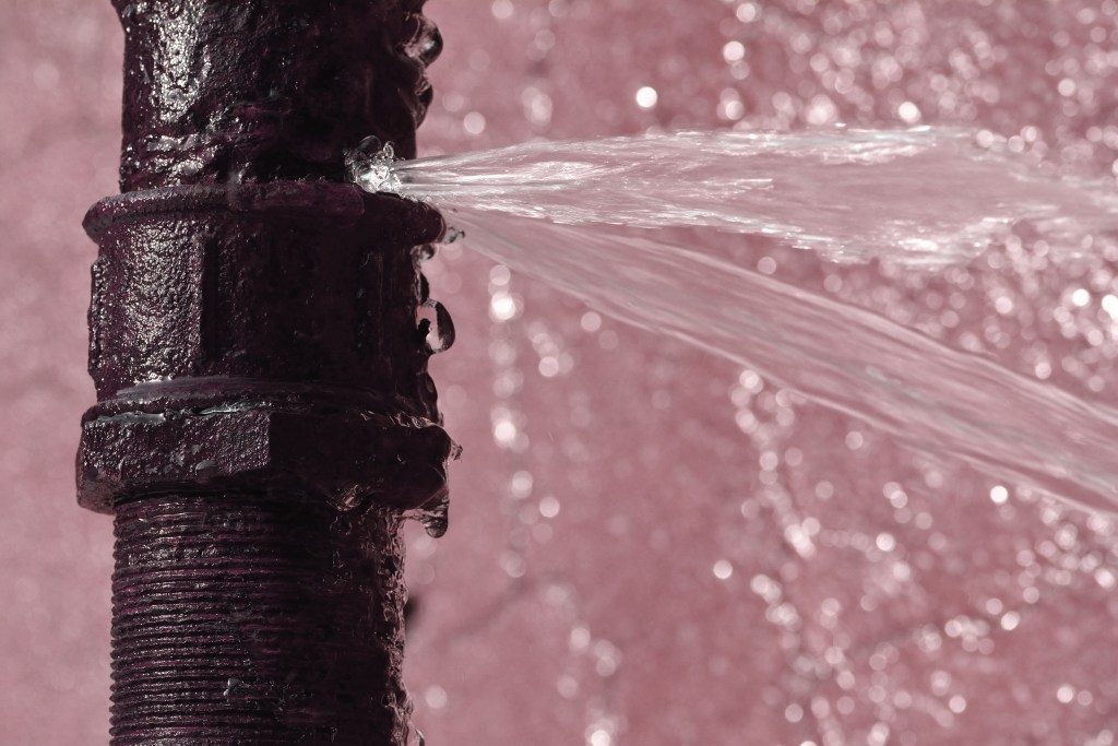 an old rusty pipe burst with water spewing out with a light pinkish purple background