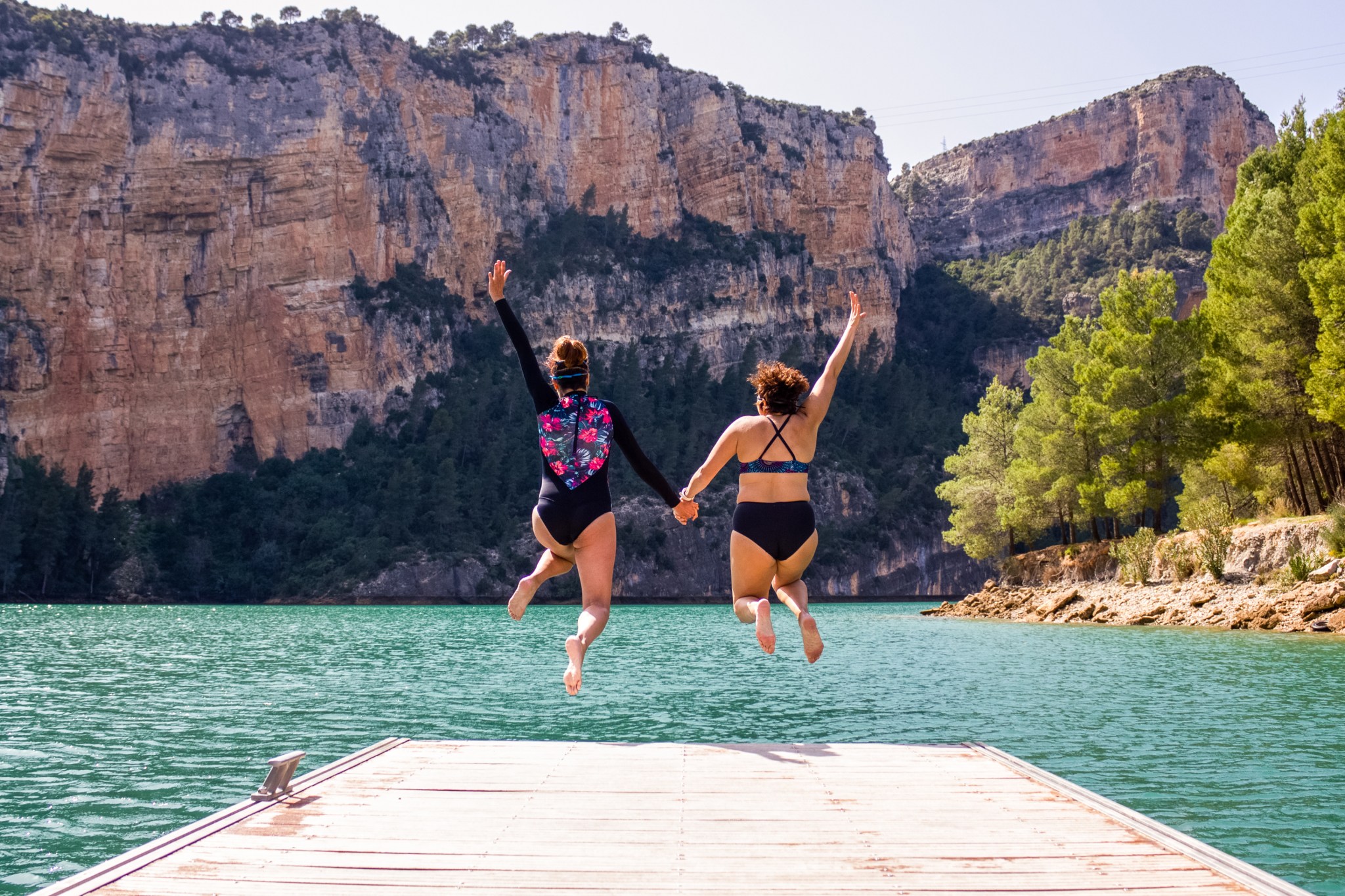 rear view of two women in bathing suits holding hands while jumping into the water from a jetty