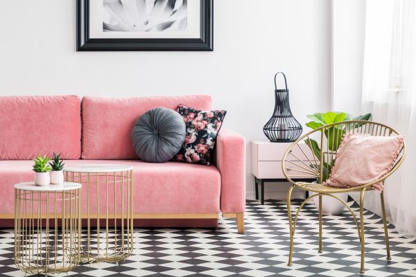 photo of a living room interior with a pink velvet sofa golden armchair and tables with decorative lights and plants with a wall painting and checkered tile floor