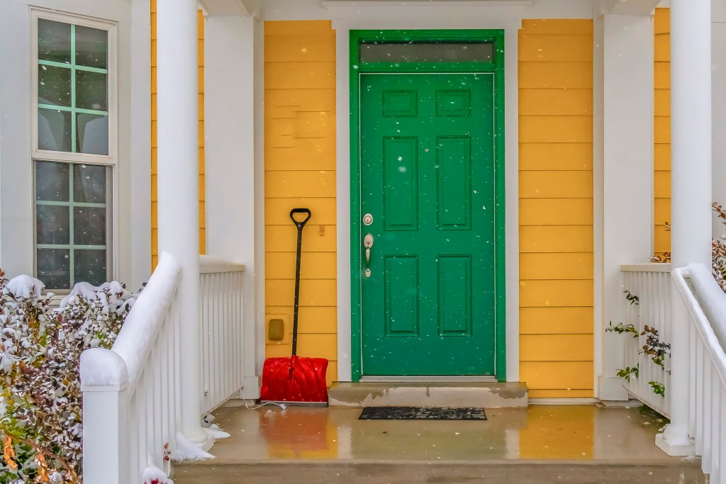 a red snow shovel leaning on the yellow siding beside the green front door of a home with white columns and a wet porch and stairs from the melting snow and there are snowflakes blurred in the foreground of the image