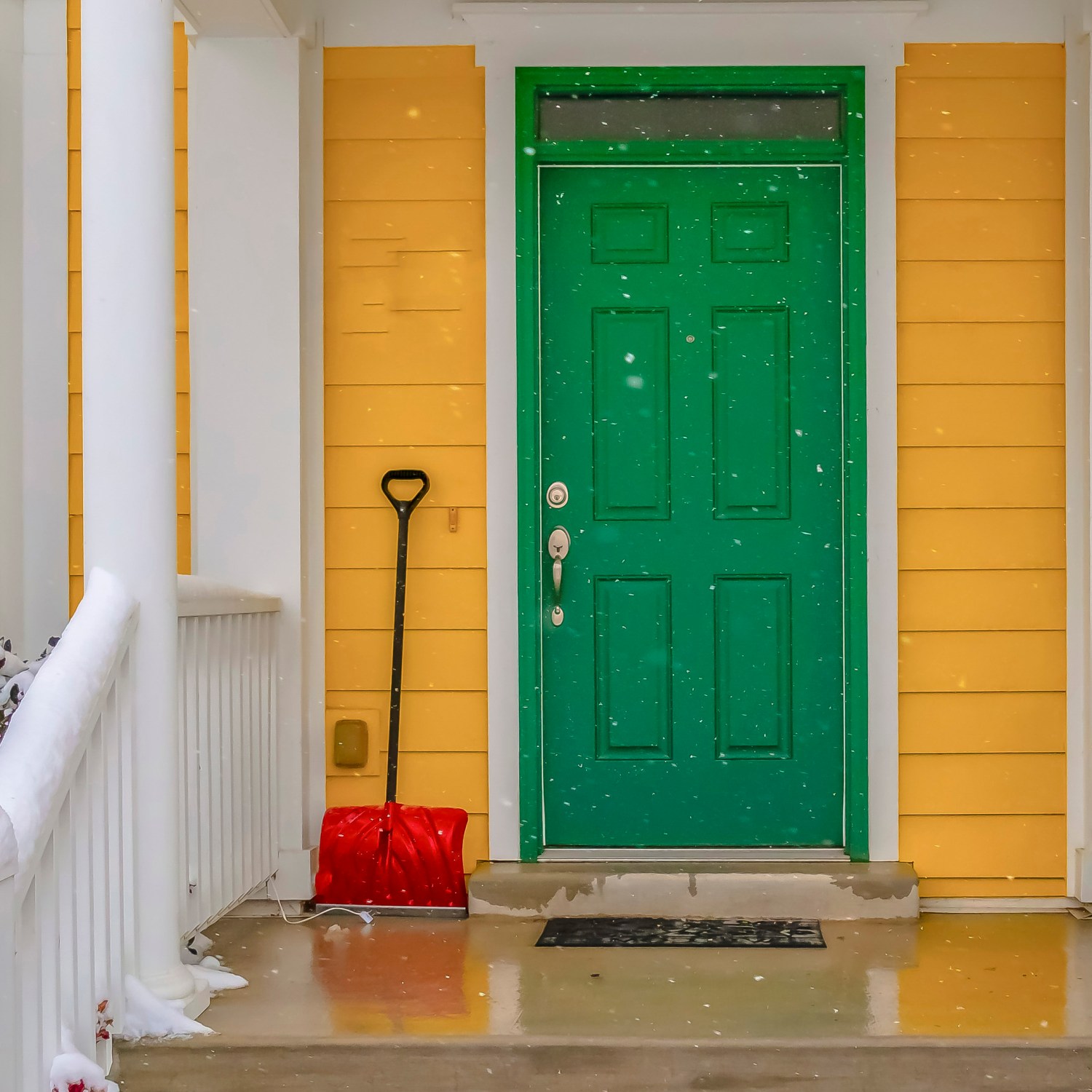 a red snow shovel leaning on the yellow siding beside the green front door of a home with white columns and a wet porch and stairs from the melting snow and there are snowflakes blurred in the foreground of the image