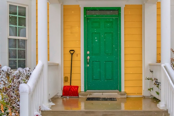 a red snow shovel leaning on the yellow siding beside the green front door of a home with white columns and a wet porch and stairs from the melting snow and there are snowflakes blurred in the foreground of the image