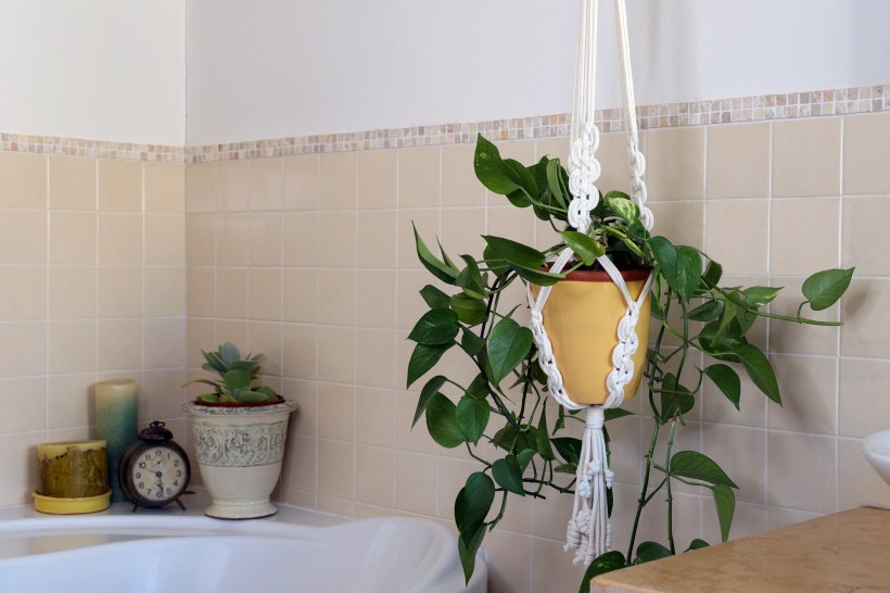 white macrame plant hanger with yellow pot holding green plant next to tub and with candles in bathroom