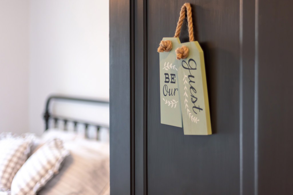 be a good home host with view into guest bedroom from an open door with a sign that says be our guest