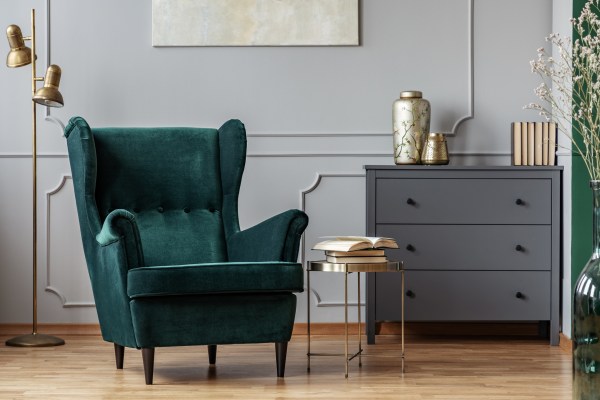 trendy home design of small golden table next to green velvet wing back chair in gray living room
