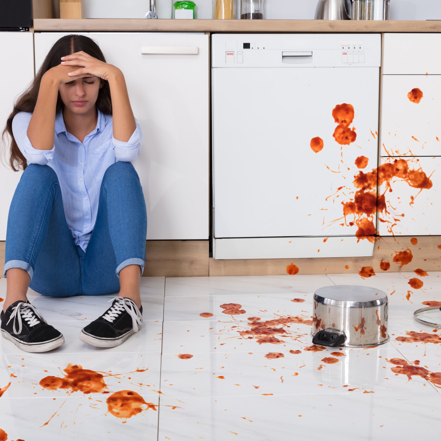 unhappy woman sitting on white kitchen floor with spilled food on the floor and white kitchen cabients