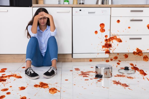 unhappy woman sitting on white kitchen floor with spilled food on the floor and white kitchen cabients