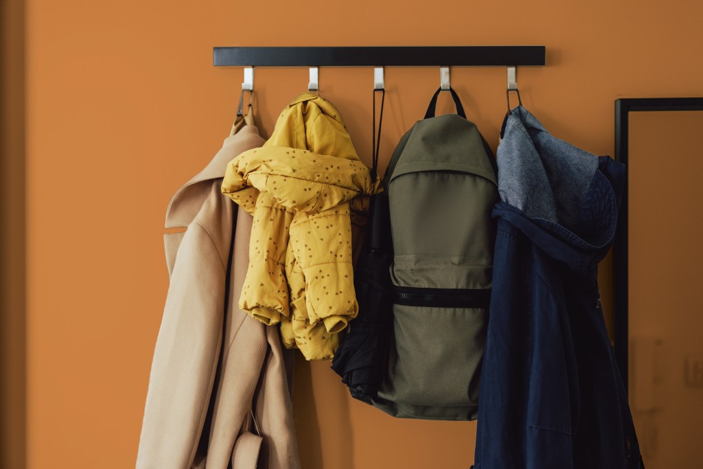 organized coatrack with three jackets and a backpack hanging on coathook on orange wall in a decluttered home