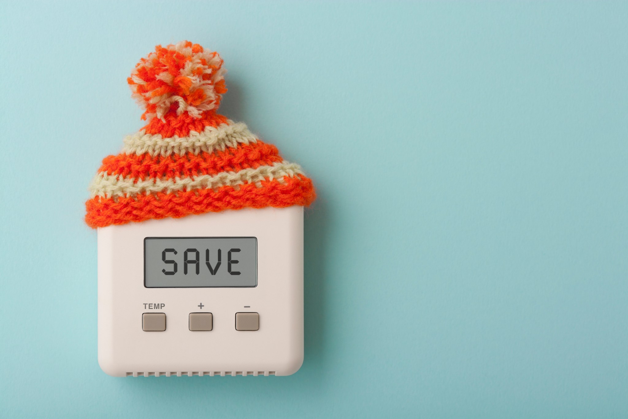 the word SAVE on a digital thermostat wearing wooly knit hat on a light blue wall for heating cooling