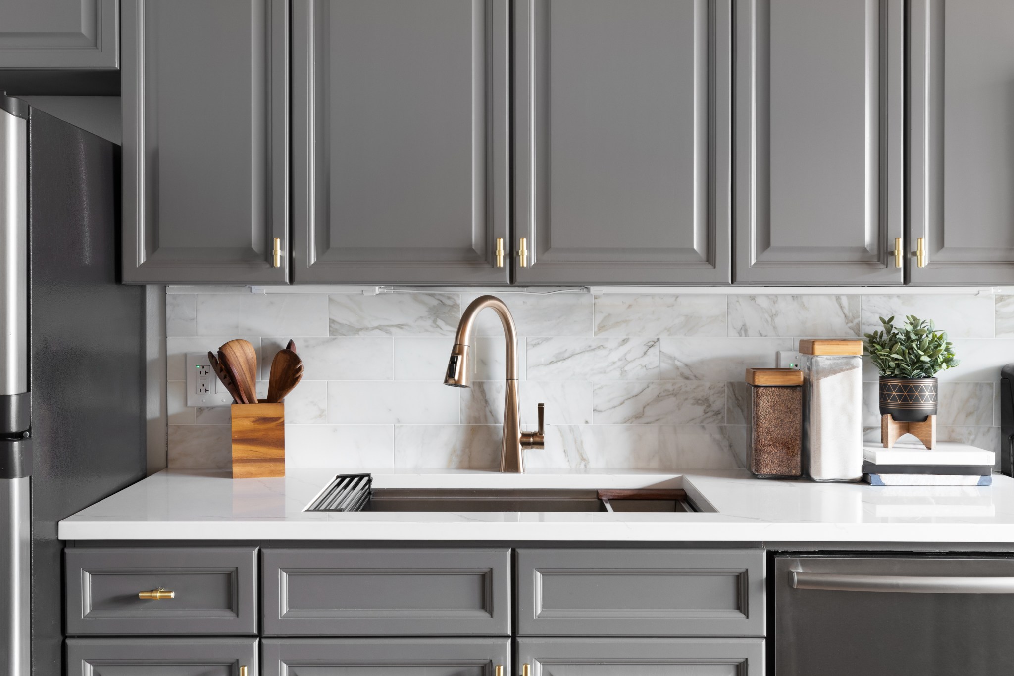 A remodeled kitchen sink detailed with grey cabinets, a white marble countertop and backsplash, and decorations.