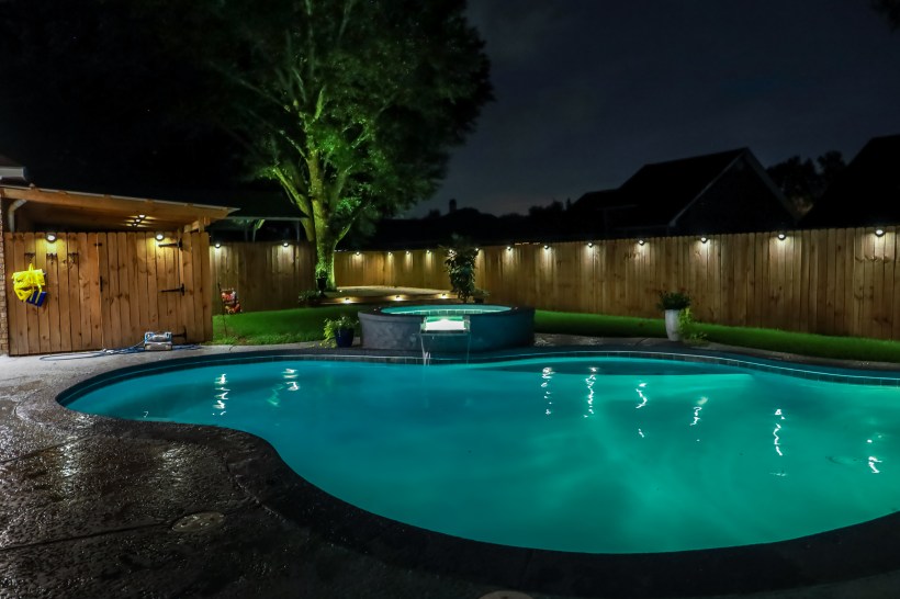 A backyard swimming pool and hot tub at night with lights around the fence.