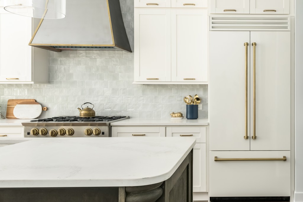 Elegant kitchen with a timeless white design, this kitchen features a white matte refrigerator, white cabinets, and two toned kitchen island with a gray tile backsplash.