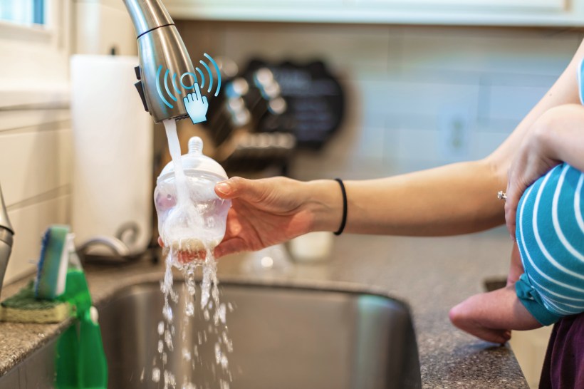 A mother using a touch activated faucet to rinse a baby bottle while holding her child.