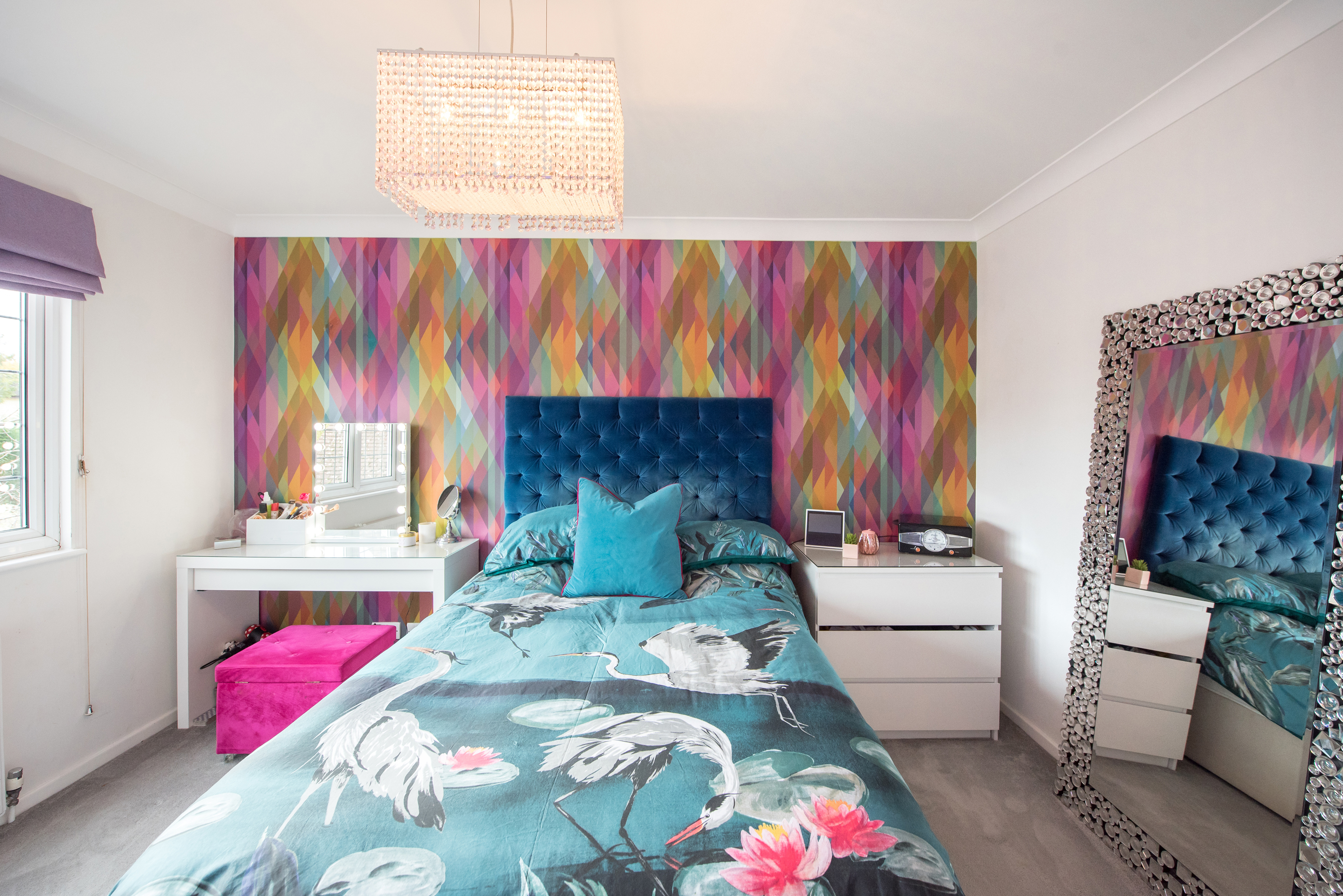 A general interior view of a bedroom, painted white with one wall decorated with a geometrical multicoloured patterned wallpapered, teal buttoned velvet headboard, double bed with floral and Heron bird patterned duvet, large crystal leaner mirror, dressing table with Hollywood makeup vanity light up mirror, chest of drawers, radio and chandelier