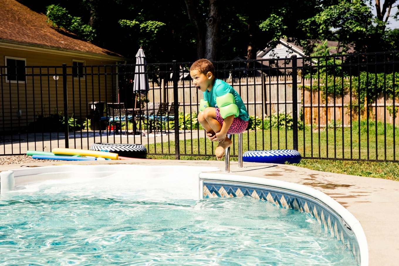 A boy jumping into a swimming pool with a bar style fence.