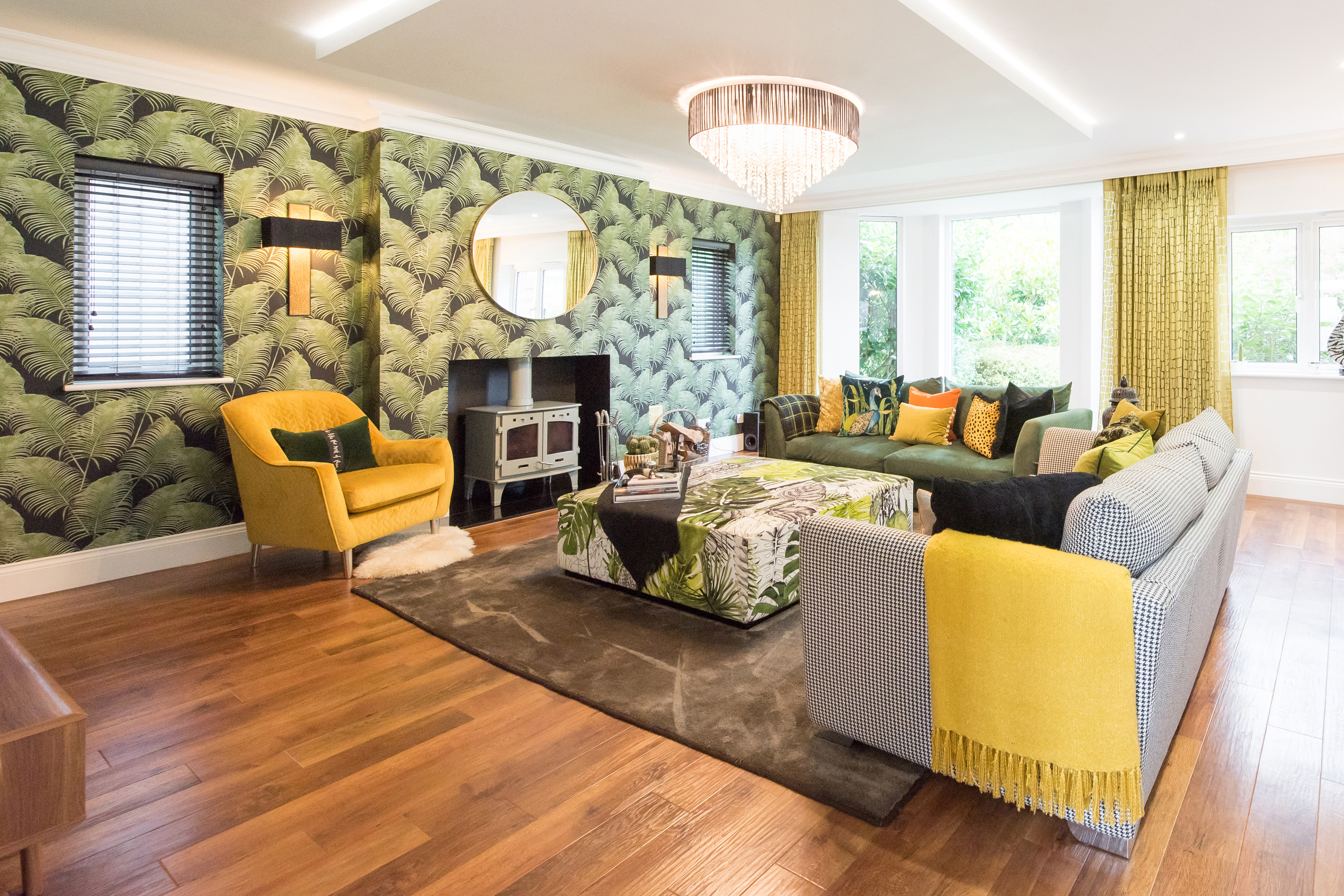 A general interior view of a large lounge with tropical palm leaf print wallpaper, wood burner stove fireplace, two sofas, armchair, chandelier, coffee table, dropped ceiling box with LED lighting, rug, hardwood flooring, black and gold wall lights.