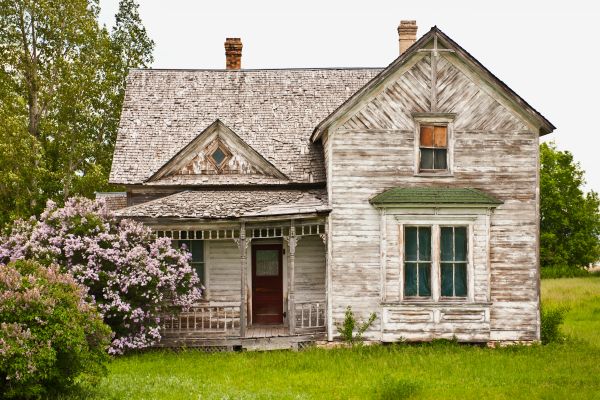 A dilapidated, abandoned fixer-upper house with lots of visible problems sitting in a nice yard with beautiful trees.